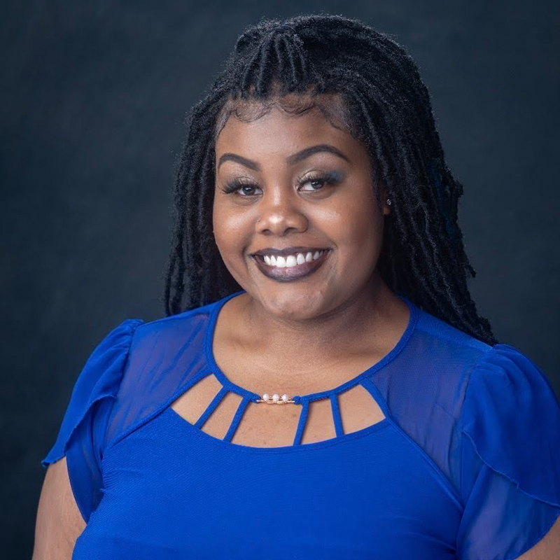 young african american woman with bright blue blouse and makeup done has her headshot taken in front of a navy backdrop