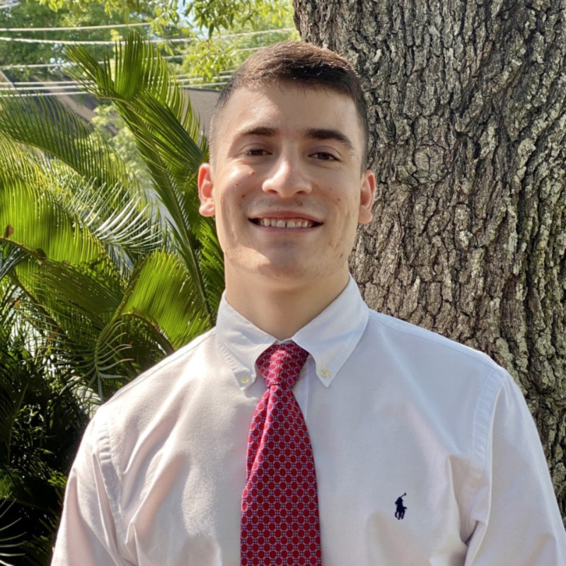 A caucasian male with short brown hair stands in front of a tree trunk and exotic greenery. He is wearing a white Polo button down with a red patterned tie.